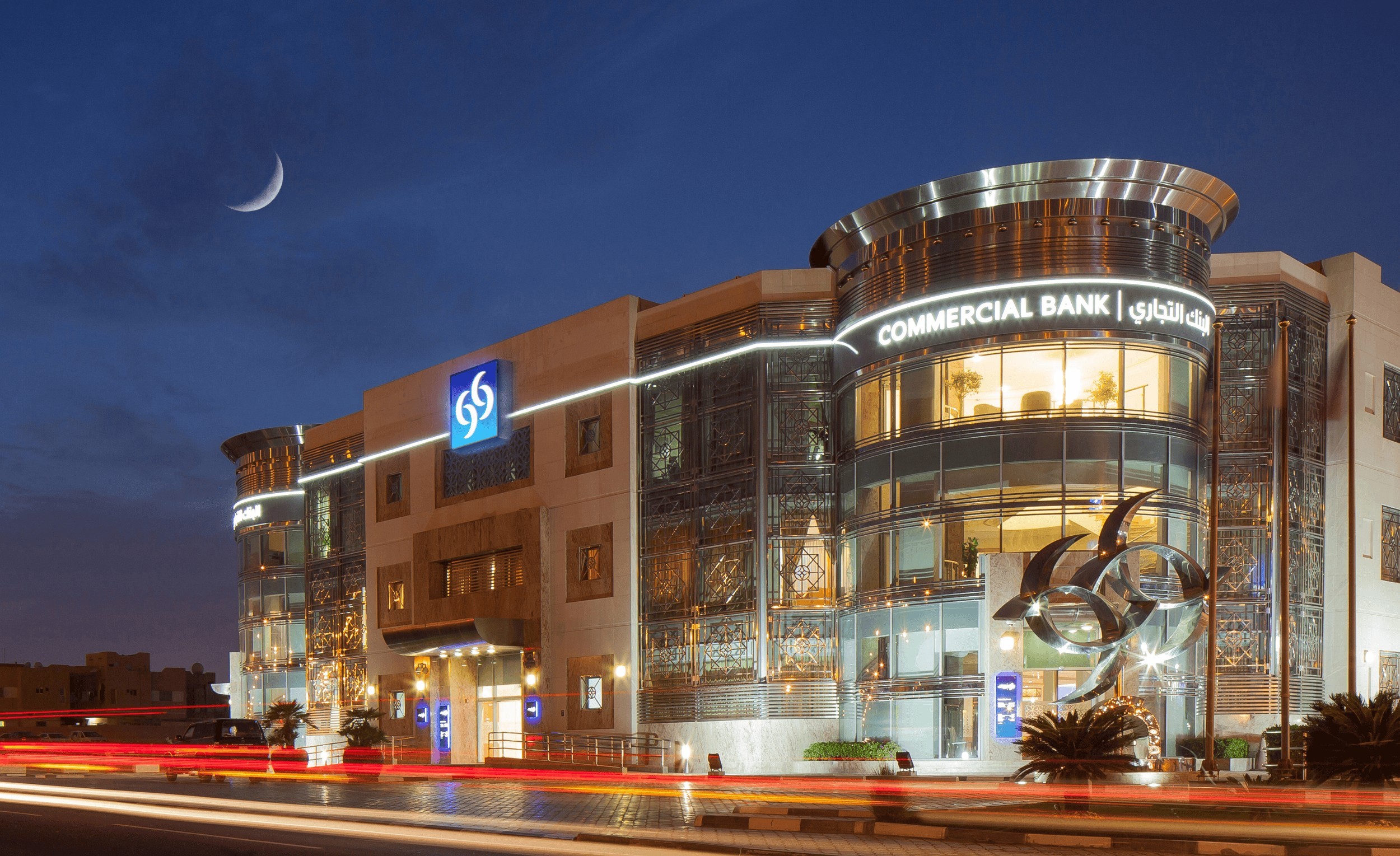 Commercial Bank retail branch in Doha at dusk with the illuminated four-crescent symbol and bilingual signage